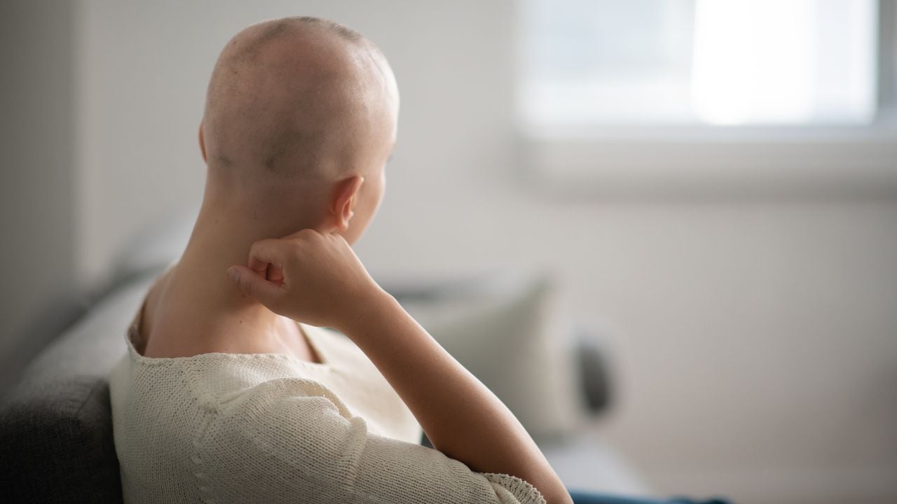 Una mujer está adentro en su sala de estar. Su cabeza está rapada debido a la quimioterapia. Ella está sentada y mirando pensativa