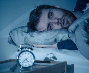Sleepless and desperate young caucasian man awake at night not able to sleep, feeling frustrated and worried looking at clock suffering from insomnia in stress and sleeping disorder concept.