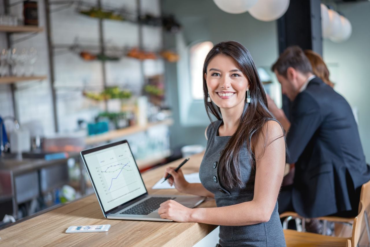 Mujer de negocios feliz trabajando en un café en una computadora portátil y mirando a la cámara sonriendo. La imagen en pantalla es de diseño propio.