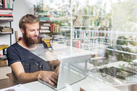 Young bearded man sitting on desk in his home office and working on laptop.