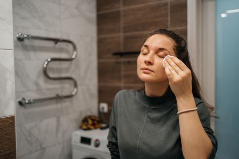 woman wiping face with moisturizing lotion with cotton pad in front of mirror in bathroom at home