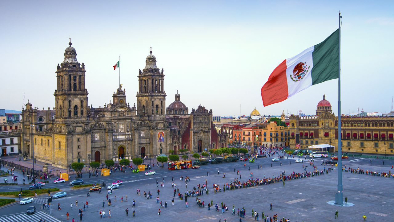La bandera mexicana ondea sobre el Zócalo, la plaza principal de la Ciudad de México. La Catedral Metropolitana está frente a la plaza, también conocida como Plaza de la Constitución.