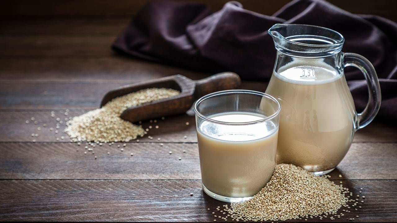 Vista frontal de una jarra y un vaso lleno de leche de quinua. El vaso y la jarra están en el lado derecho de la imagen sobre una mesa rústica de madera, al lado del vaso hay un montón de semillas de quinua. Esta imagen es parte de una serie de leches veganas.