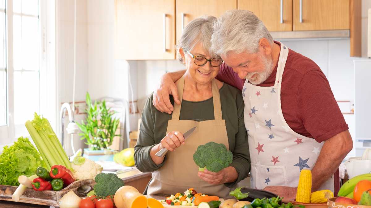 Vegetarian lifestyle. Beautiful smiling senior couple white-haired hug in the kitchen preparing a vegetable soup. On the table a mix of raw seasonal vegetables and a broccoli in the hand