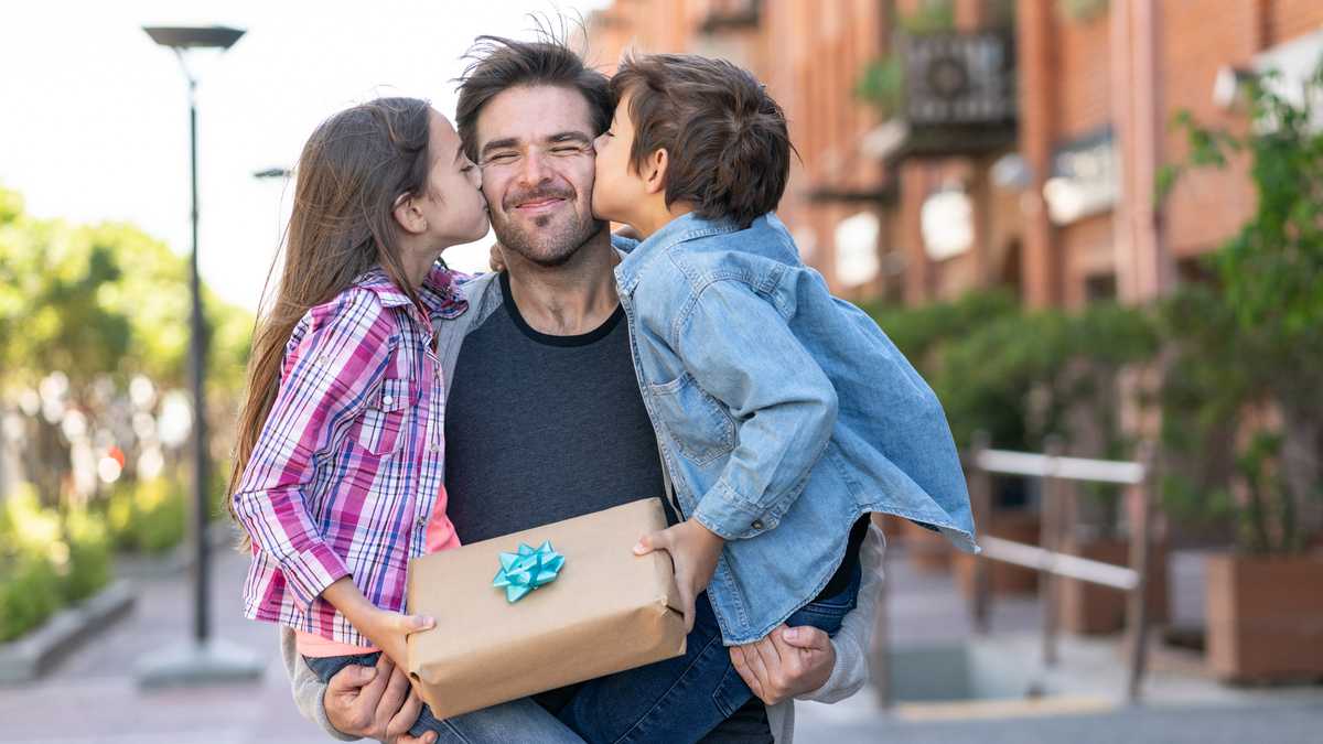 Amoroso hijo e hija besando a papá en la mejilla mientras él los lleva a ambos con un regalo para el día del padre - Conceptos de celebración