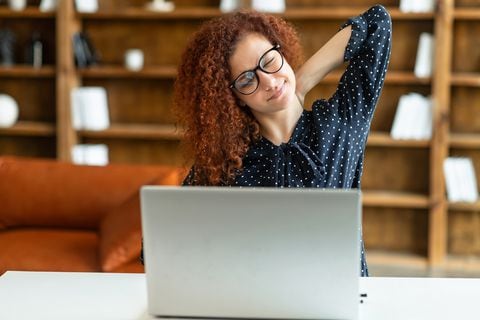 Mujer despreocupada atractiva, mujer rizada pelirroja en gafas que estira los brazos sentados en el escritorio en la oficina, la empresaria sofisticada descansa después de un trabajo bien hecho computador