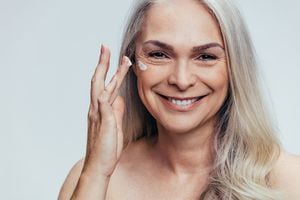 Sonriente mujer caucásica de mediana edad aplicando crema antienvejecimiento en la cara. Mujer mayor aplicando crema hidratante en la cara contra un fondo gris.