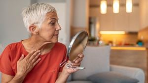 Hermosa anciana de pelo corto sosteniendo un espejo y aplicando crema facial en casa. Atractiva anciana de pelo gris mirándose en el espejo mientras se sienta en el sofá durante el día.