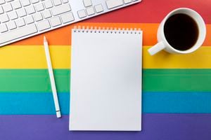 Bloc de notas, bolígrafo blanco, taza de café y teclado sobre fondo despojado de colores de la bandera del arco iris que simbolizan el movimiento LGBT.