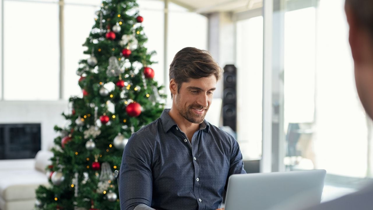 Happy young businessman working on laptop during christmas holiday. Successful business man in modern office working with xmas tree in background. Entrepreneur using laptop in modern office.