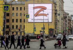 Los peatones cruzan una calle frente a una valla publicitaria que muestra el símbolo "Z" con los colores de la cinta de San Jorge y un lema que dice: "No nos damos por vencidos con nuestro pueblo", en apoyo de las fuerzas armadas rusas, en San Petersburgo, 7 de marzo de 2022 (Foto por AFP)
