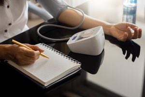 Mujer tomando presión arterial usando esfigmomanómetro digital y grabando en un cuaderno o diario blanco vacío en casa. Concepto médico y sanitario.