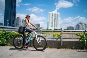 Vista lateral feliz sonriente mujer de negocios de 45 años en bicicleta en bicicleta eléctrica al aire libre en un día soleado de verano en la ciudad europea, edificios de oficinas borrosa en el fondo
