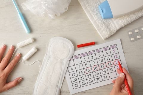 Woman marking critical menstrual days on calendar with pills and intimate care around on white wooden table. Top view.