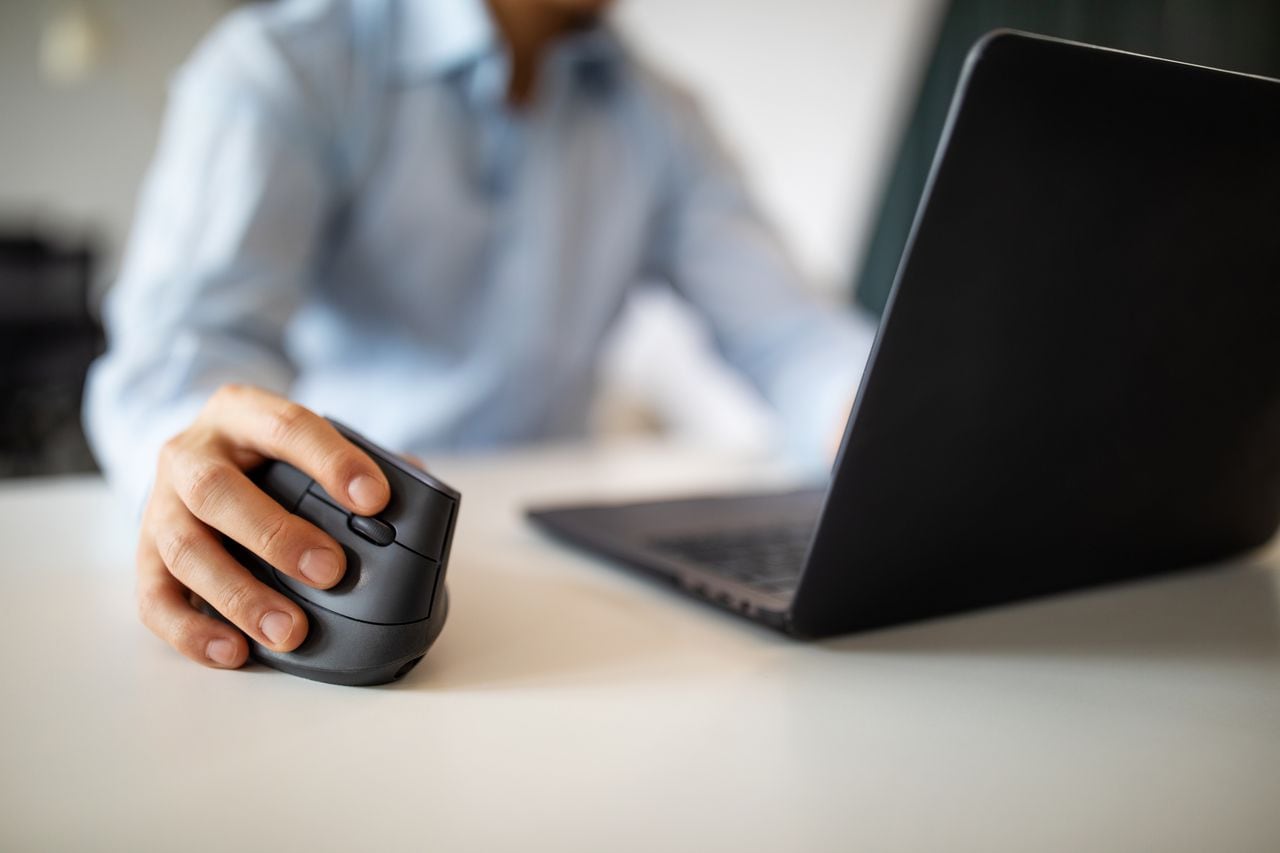 Close up of man using computer mouse while working on a laptop in office. Focus on male hand using computer mouse on desk.