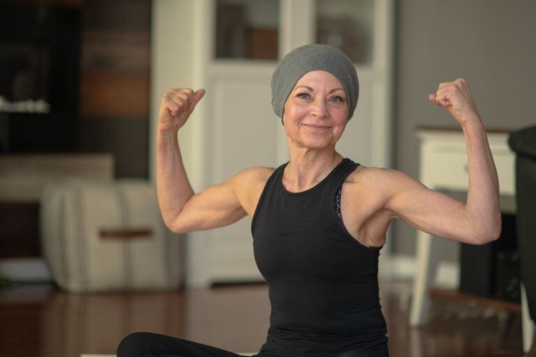 Una mujer mayor con cáncer flexiona sus músculos mientras está sentada en su piso en su casa. Ella sonríe porque se está volviendo más fuerte.