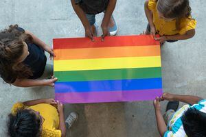 A group of multi-ethnic elementary students stand around a colorful gay pride poster.  They are each holding an edge of the poster so it is out flat for the camera to see.  Only the tops of their heads can be seen as the picture is taken from an aerial view.  Each of the students are wearing casual clothing.