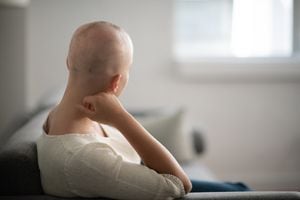 Una mujer está adentro en su sala de estar. Su cabeza está rapada debido a la quimioterapia. Ella está sentada y mirando pensativa
