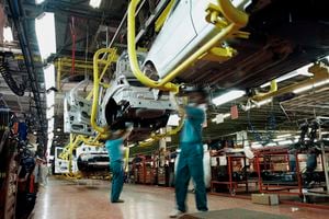 Dos trabajadores vestidos con camisas verdes y pantalones azules trabajan debajo de un automóvil en una fábrica de automóviles. Hay líneas de montaje, automóviles parcialmente construidos, herramientas y equipos eléctricos al fondo.