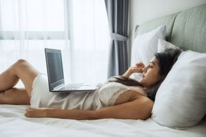 Hermosa mujer bronceada asiática sexy en pijama blanco mira películas en línea desde una computadora portátil en un dormitorio moderno. Chica relajarse en la cama en la mañana del fin de semana. modelo web webcam