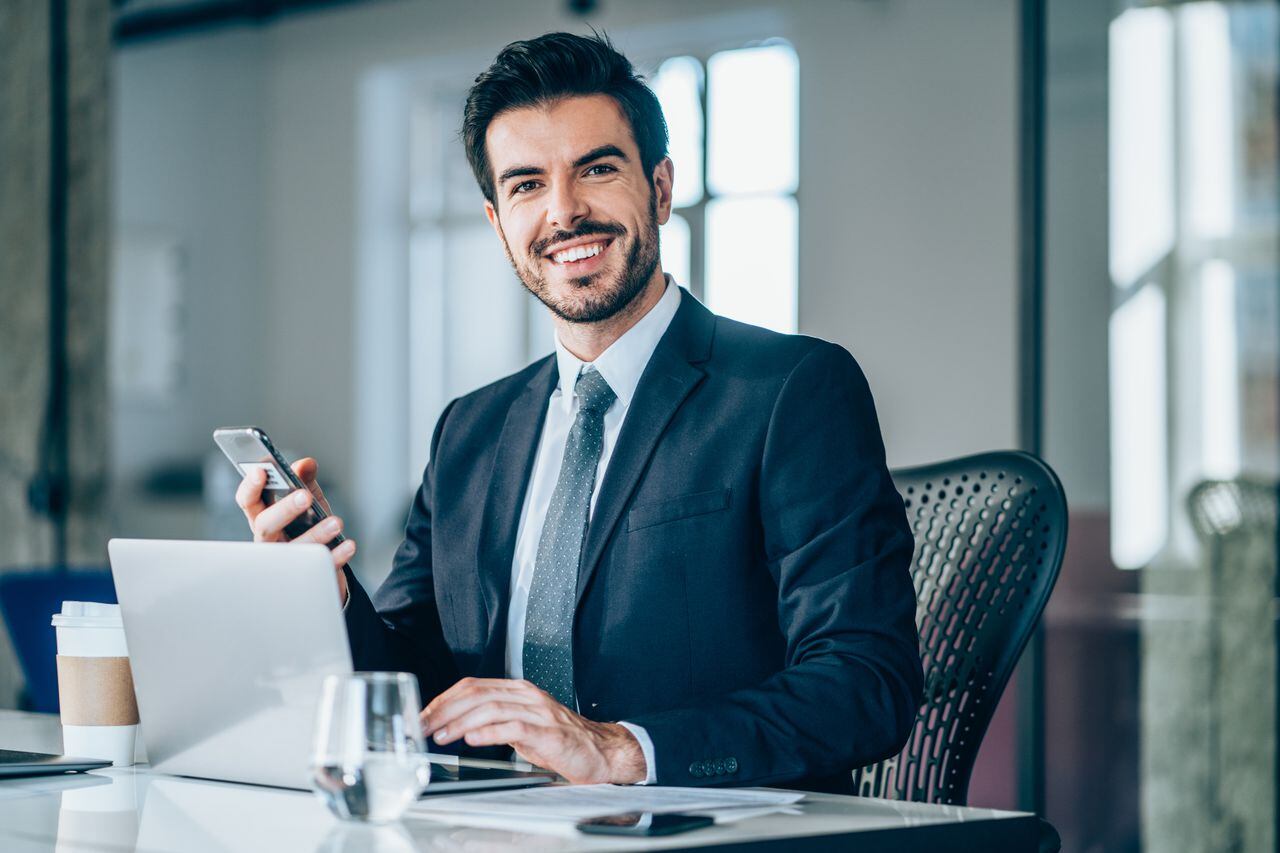 Foto de un joven empresario sentado en el escritorio en la oficina moderna y mirando a la cámara. Retrato de un apuesto hombre de negocios trabajando en equipo portátil y usando teléfonos inteligentes en la oficina.