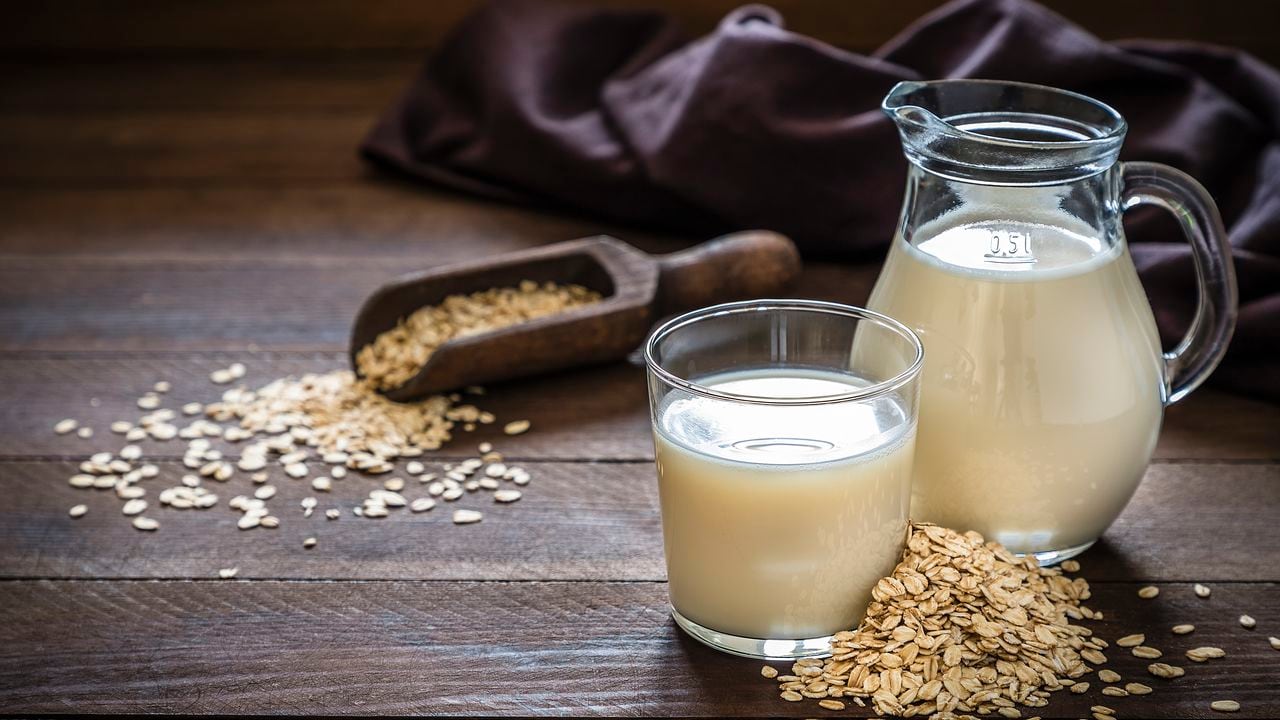 Front view of a jug and a drinking glass full of oat milk. The glass and the jug are at the right side of the image on a rustic wooden table, next to the glass its a heap of oat flakes. This image is a part of a vegan milk series. Low key DSLR photo taken with Canon EOS 6D Mark II and Canon EF 24-105 mm f/4L