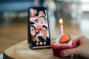 Close-up shot of a birthday cake and a smart phone screen showing international friends having a virtual birthday party on video call.