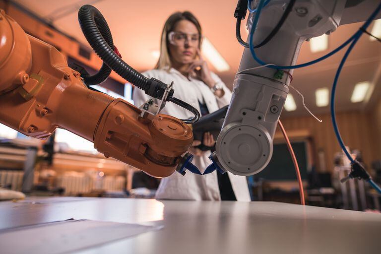 Dos brazos robóticos que fabrican algo en el laboratorio mientras una ingeniera está en el fondo