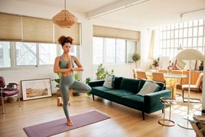 Full length of fit woman practicing tree pose at home. Young woman standing on one leg on exercise mat. She is doing yoga in living room.
