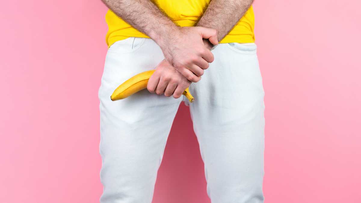 Potency and men's health. A man in white jeans, legs apart, holds a banana near the genitals. Pink background. Close up of hands.
