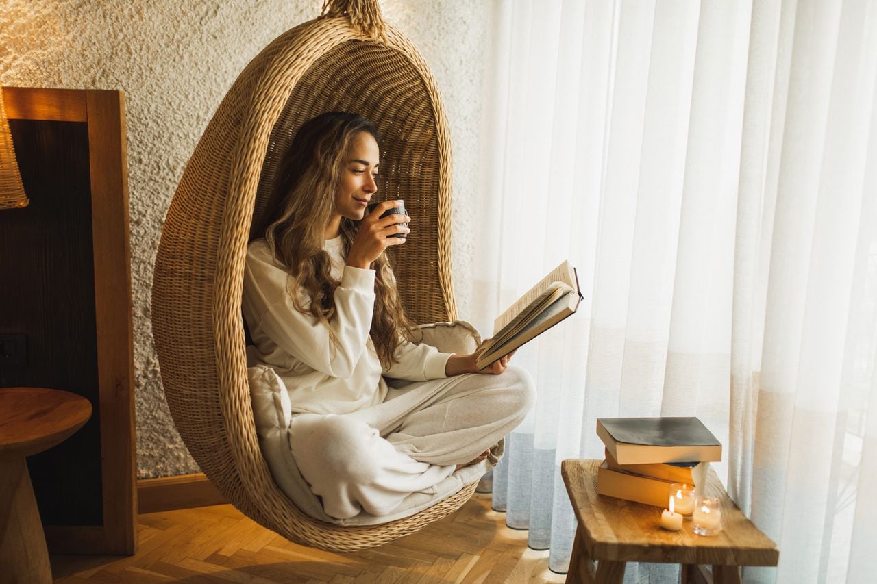 Wellbeing and rest. Young attractive brunette woman with long hair sitting in wicker egg swing chair. Leisure and life balance. Reading as hobby. Coziness and natural wooden furniture at home, burning candles, sustainable living.