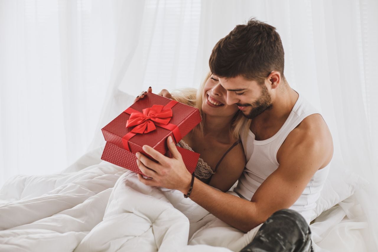 Front view of young couple getting up from bed in a white room. Woman is surprised by her couple bringing a red gift box during their anniversary celebration.