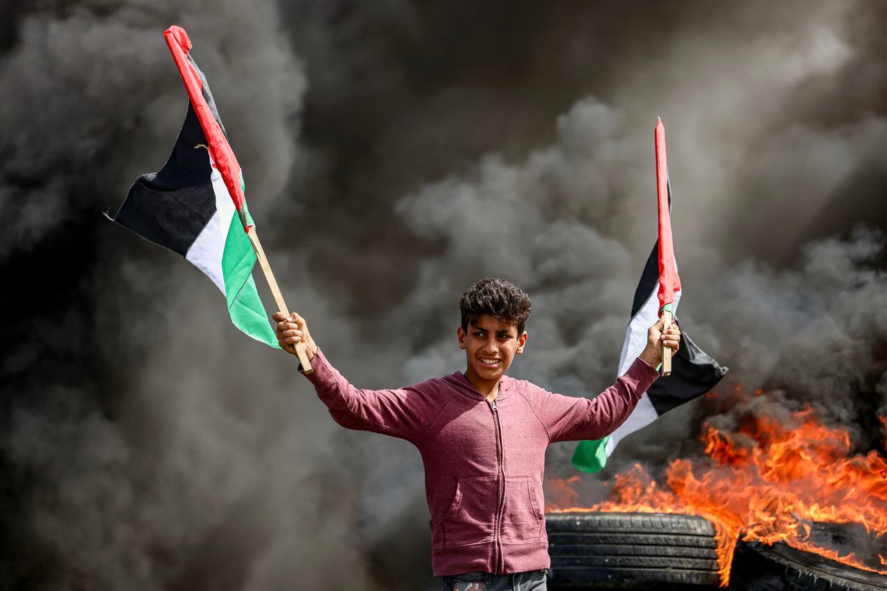 Un joven sostiene banderas palestinas junto a los gases de los neumáticos en llamas durante una manifestación a lo largo de la frontera con Israel al este de la ciudad de Gaza el 5 de abril de 2023. 
 (Foto de MAHMUD JAMONES / AFP)