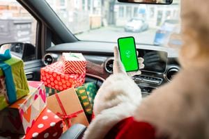 A medium, over-the-shoulder shot of a senior man wearing a Santa Claus costume. He sits in his delivery van with a stack of Christmas presents on the seat beside him. He is holding his smartphone showing a graphic which reads "Payment Sent" with a tick above.