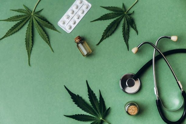 Flat lay ripe cannabis leaves, white pack of pills, stethoscope and hemp oil jars lying down on green background.
