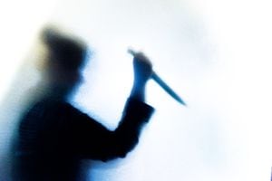 Backlit image of the silhouette of a person, probably a woman, wielding a sharp knife in an aggressive way. The silhouette is distorted, and the arms elongated, giving an alien-like quality. The image is sinister and foreboding, with an element of horror. The image conveys a domestic violence, knife crime theme. Horizontal image with copy space.
