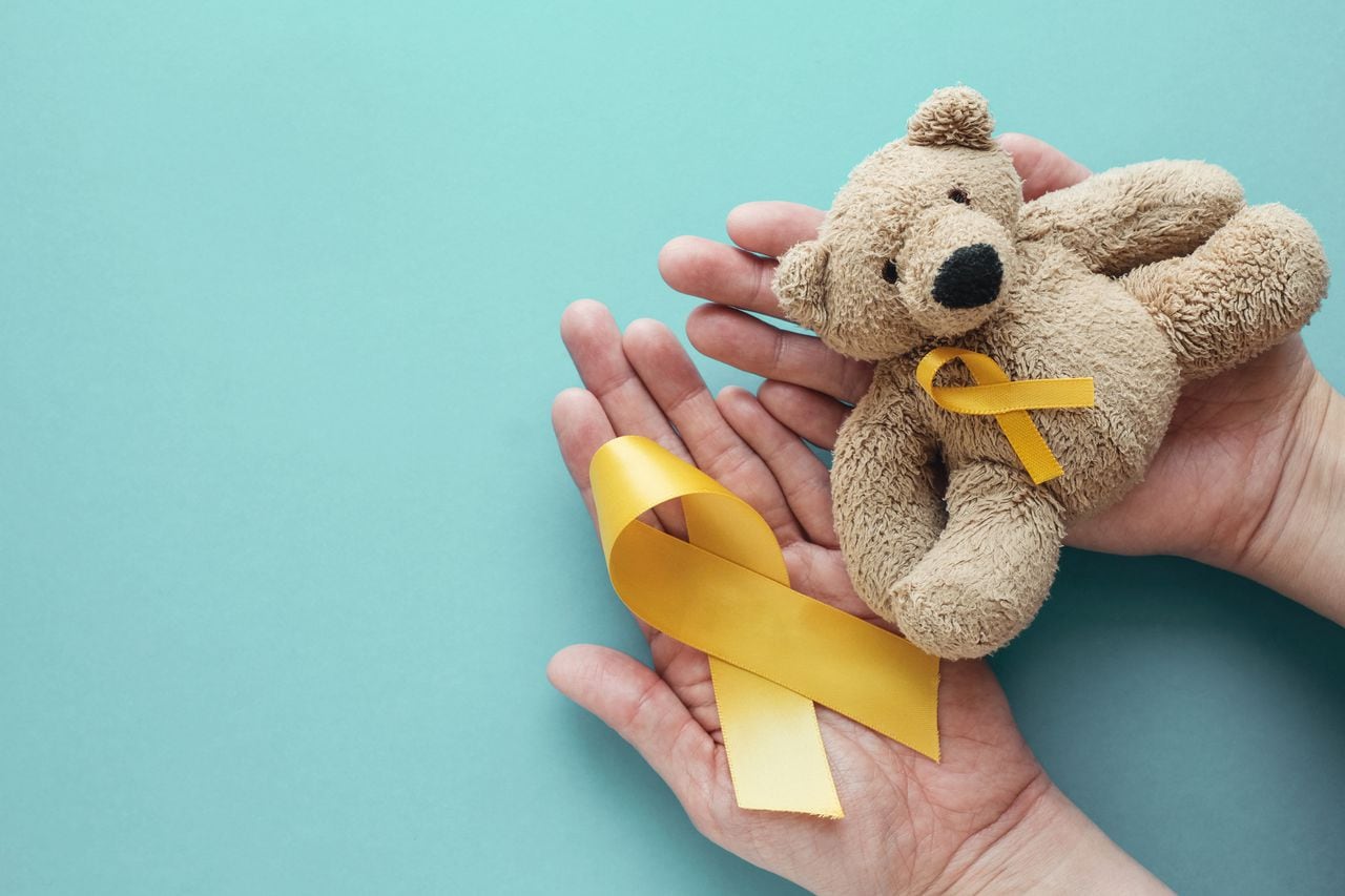 Manos sosteniendo a los niños oso pardo de juguete suave con cinta de oro amarillo, conciencia del cáncer infantil