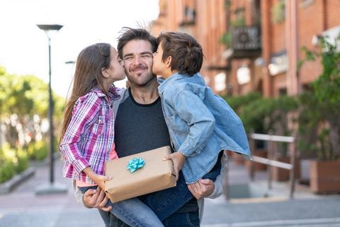 Amoroso hijo e hija besando a papá en la mejilla mientras él los lleva a ambos con un regalo para el día del padre - Conceptos de celebración