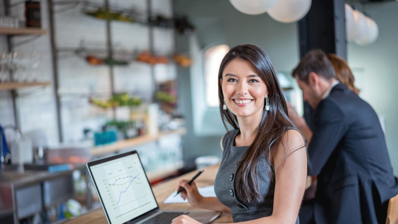 Mujer de negocios feliz trabajando en un café en una computadora portátil y mirando a la cámara sonriendo. La imagen en pantalla es de diseño propio.