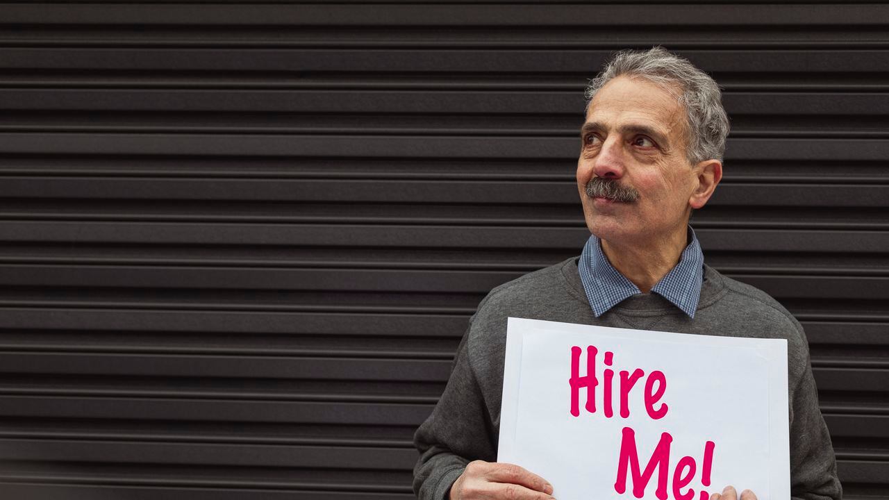 Senior asian man standing looking away from the camera while holding a hire me sign.