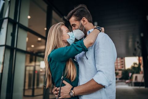 Una joven pareja con tapabocas y besándose al aire libre en la ciudad.