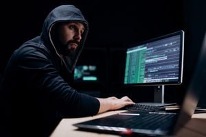 Focused computer specialist in black hoodie using computers for organizing massive data attack in internet. Bearded caucasian man working at dark underground hideout.