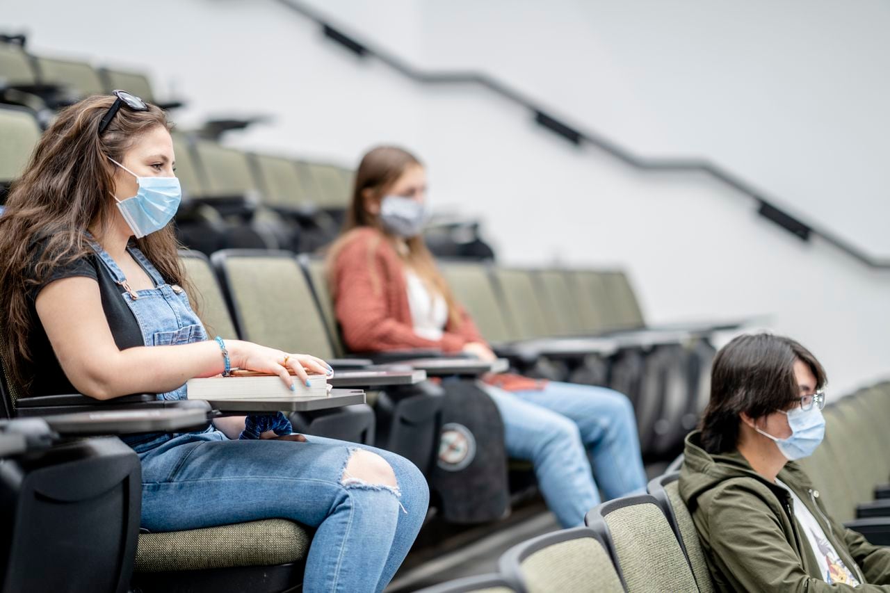 Grupo multiétnico de estudiantes que usan máscaras protectoras mientras están sentados en una sala de conferencias a 2 metros de distancia.