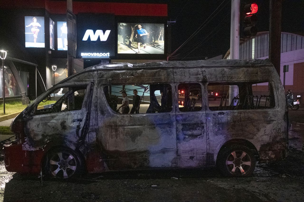 Personas observan un vehículo de transporte quemado tras ser incendiado por individuos no identificados en Tijuana, estado de Baja California, México, el 12 de agosto de 2022. Varios vehículos fueron incendiados, ese mismo día, en la ciudad. (Photo by Guillermo Arias / AFP)