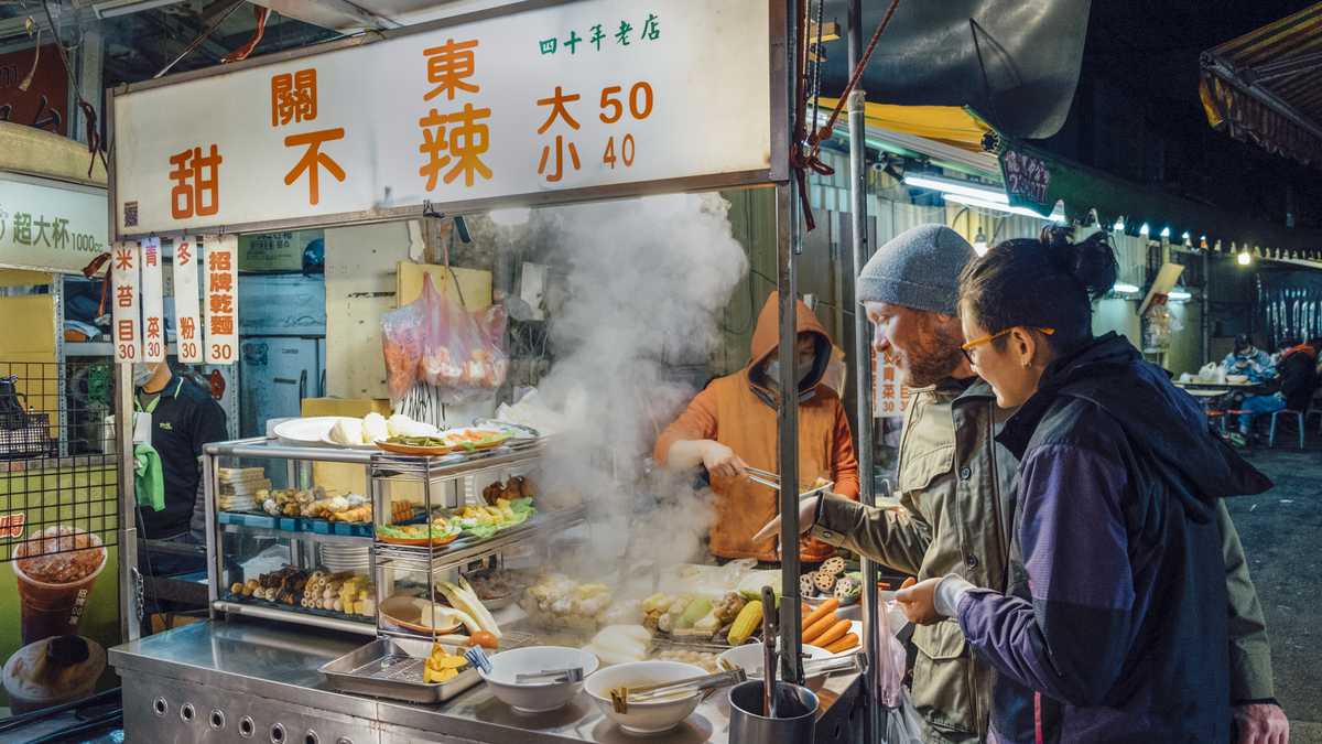 Hombres caucásicos y mujeres asiáticas en el mercado nocturno taiwanés al aire libre de pie frente al vendedor de alimentos