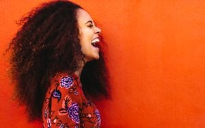 Vista lateral de una mujer alegre con el pelo rizado parado en un fondo naranja. Primer plano de una joven africana riendo.