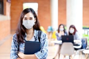 Retrato de un joven estudiante asiático con una máscara mirando la cámara sosteniendo un cuaderno o una tableta en el brazo en concepto de regreso o regreso a la escuela, reapertura de la escuela y desbloqueo después del brote de covid o coronavirus.