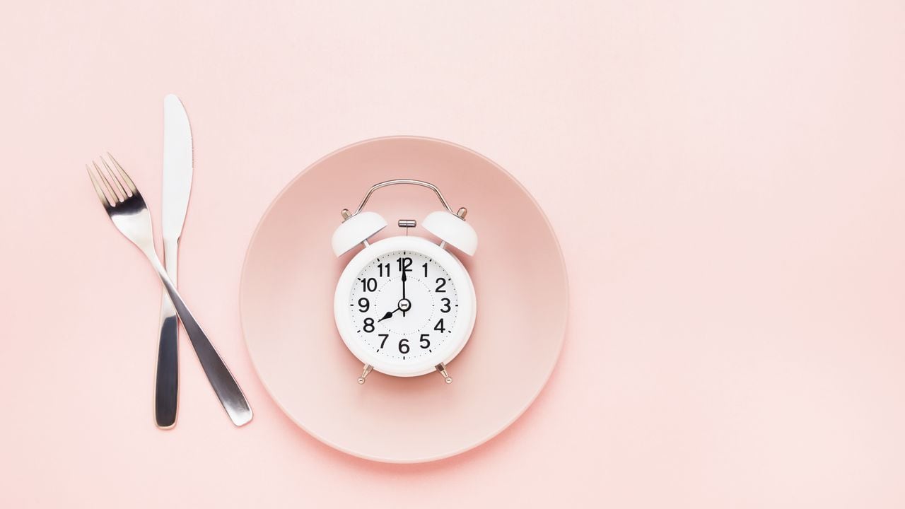 Intermittent fasting concept. White alarm clock on empty pink dish with knife and fork on pink background. Flat lay, copy space for text.