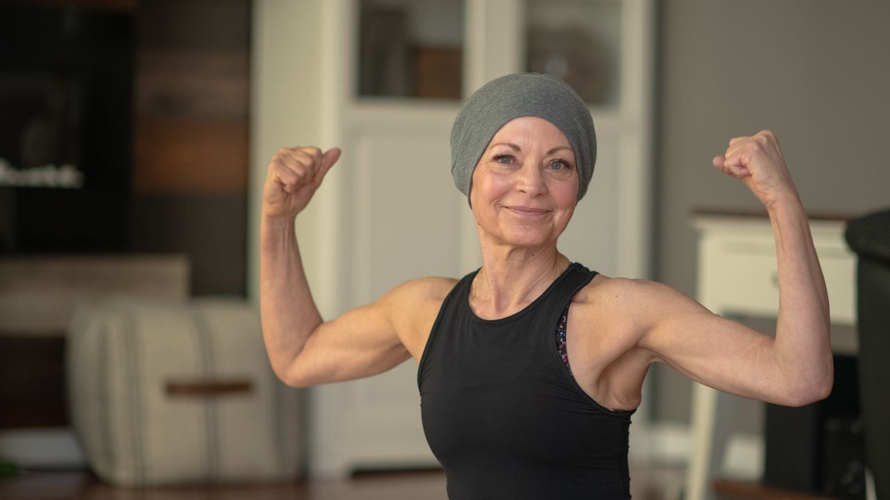 Una mujer mayor con cáncer flexiona sus músculos mientras está sentada en su piso en su casa. Ella sonríe porque se está volviendo más fuerte.
