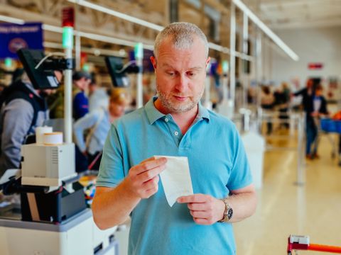 Close up color image depicting a mid adult man checking his groceries bill in the supermarket after making his purchases at the checkout. Focus on the man with the supermarket checkout defocused in the background. The man is wearing a casual blue polo shirt and has a worried expression on his face.
